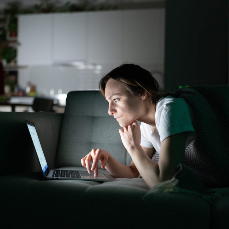 Focused young woman working online on laptop at home in the night, looking at screen, thinking, reading news media. Concentrated girl lying on sofa in dark room, using computer, suffers from insomnia